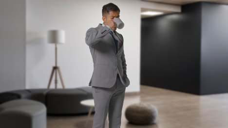 Portrait-Of-Businessman-Wearing-Suit-Standing-Drinking-Coffee-In-Modern-Open-Plan-Office